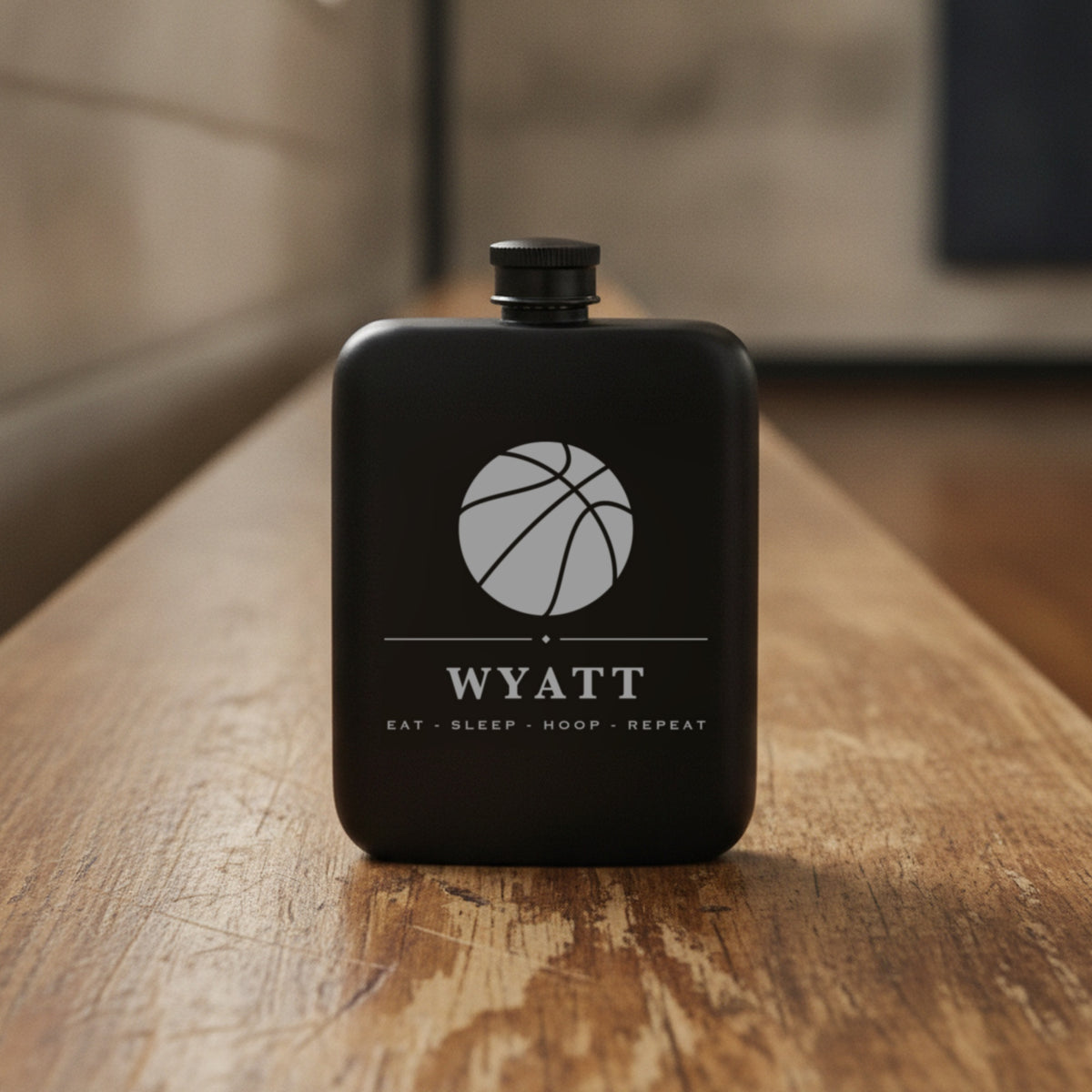 Engraved basketball groomsmen flask reading Eat Sleep Hoop Repeat standing on a worn wooden gym bench against a cinder block wall.