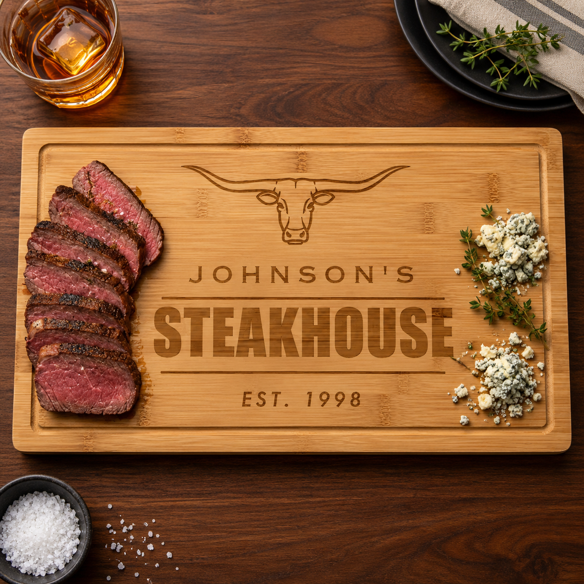 Personalized steakhouse cutting board engraved with a longhorn steer, Johnson's Steakhouse and Est. 1998, set with sliced steak, blue cheese and fresh thyme