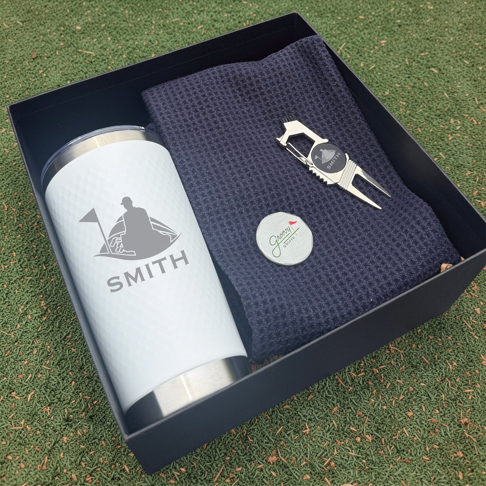 Golf-themed gift set with tumbler, towel, and divot tool in a box on grass.