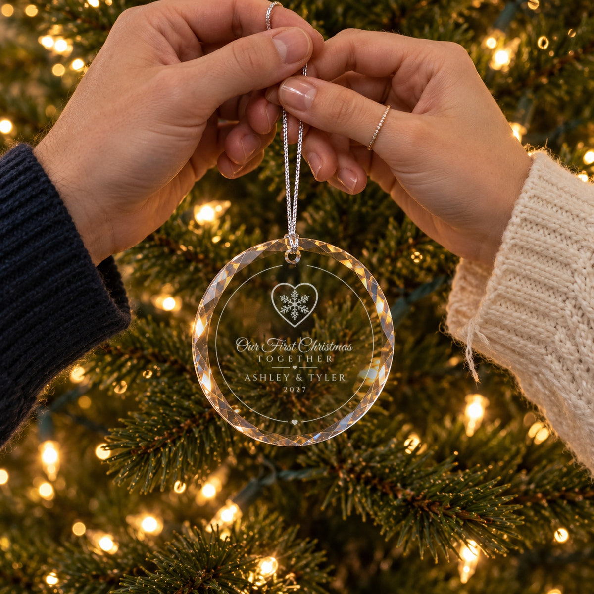 An engaged couple's hands holding the Snowflake Heart Our First Christmas Together ornament by the tree, engraved ASHLEY & TYLER, 2027