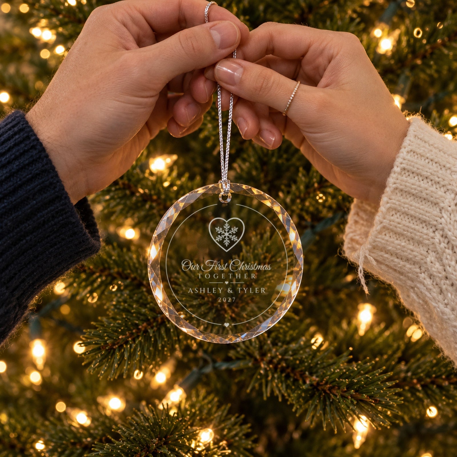 An engaged couple's hands holding the Snowflake Heart Our First Christmas Together ornament by the tree, engraved ASHLEY & TYLER, 2027