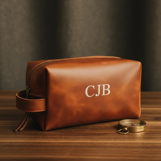 Cognac leather dopp kit with sample monogram "CJB" displayed on wooden surface next to brass compass