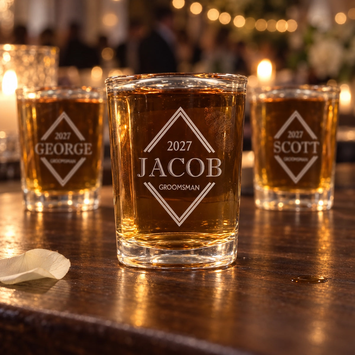 Three engraved groomsmen shot glasses filled with whiskey on a walnut bar at a candlelit wedding reception, each etched with a name, role and 2027 in a diamond.