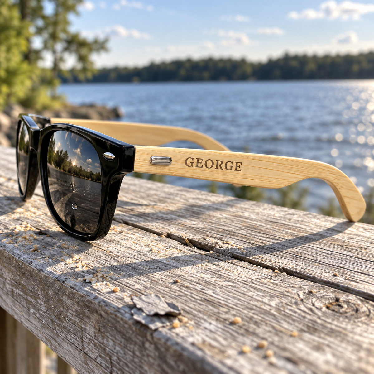 Shady Squad sunglasses with black frames lying open on a weathered deck rail, the light wood grain arm engraved GEORGE in gray-brown near the hinge