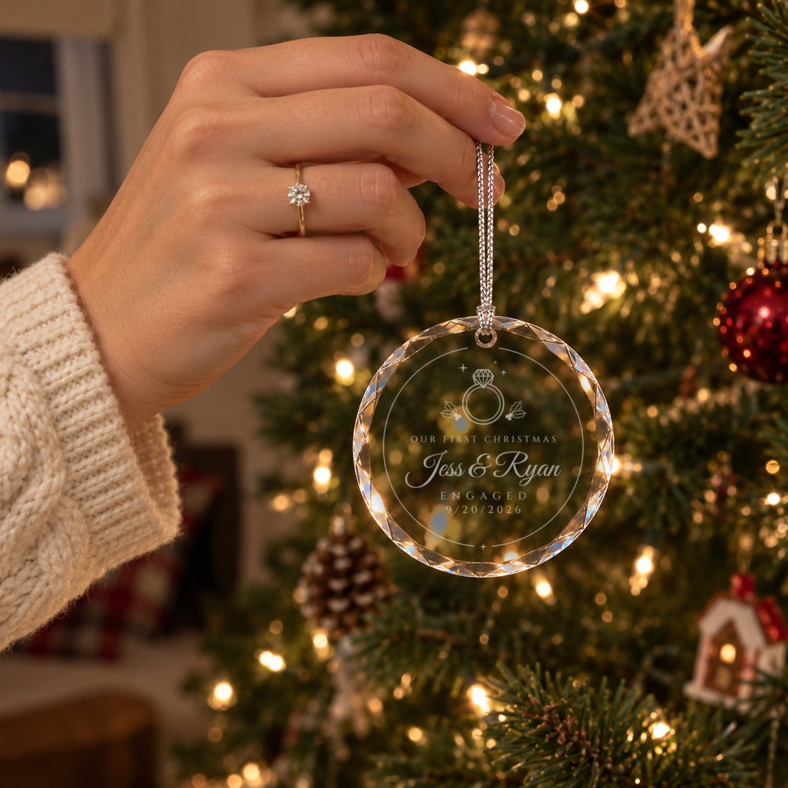 Hand wearing a solitaire engagement ring holding the first Christmas engaged ornament by its cord in front of a lit tree, engraved Jess & Ryan