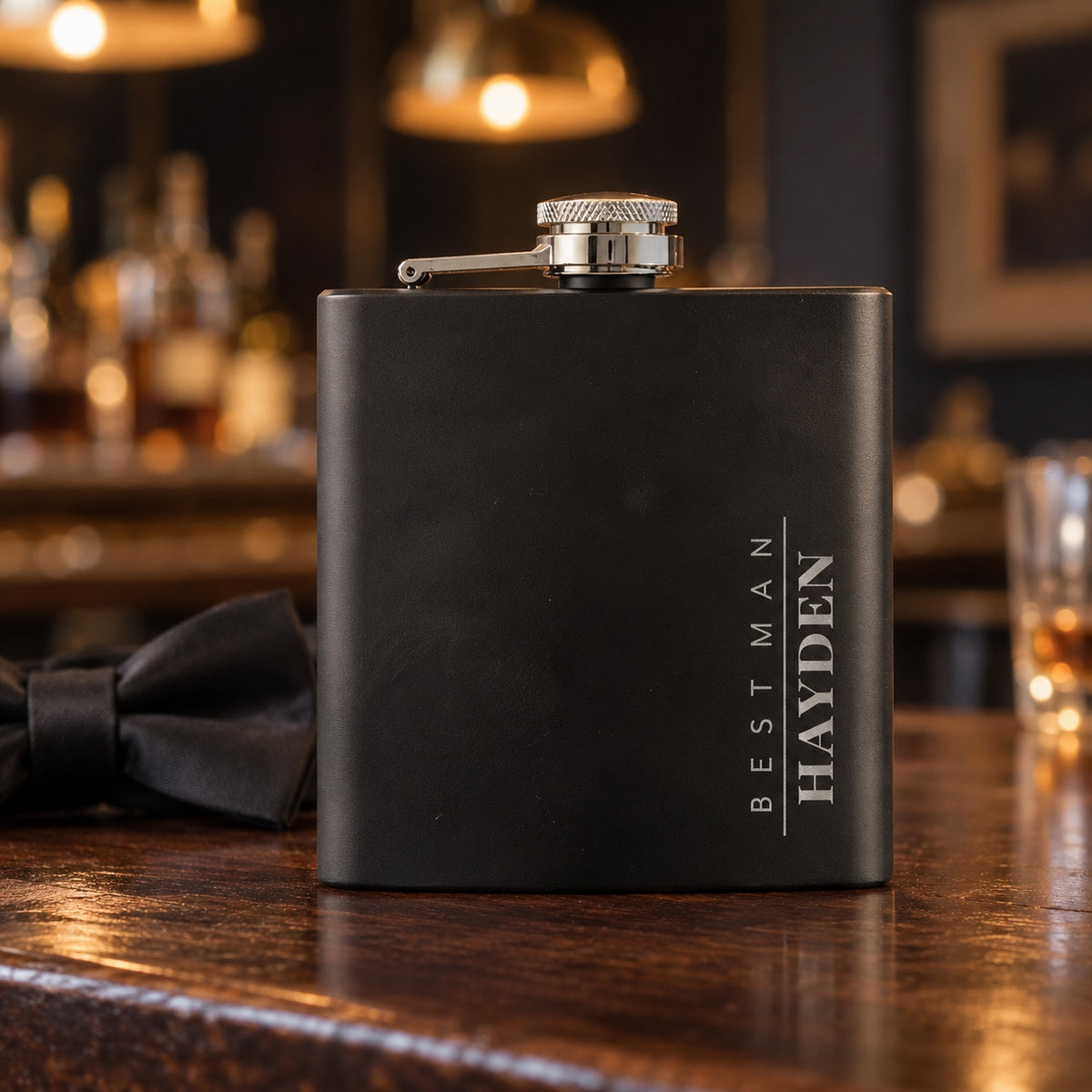 Engraved groomsmen flask in matte black on a walnut bar beside a black bow tie, the name HAYDEN running tall down the side with BEST MAN beside it