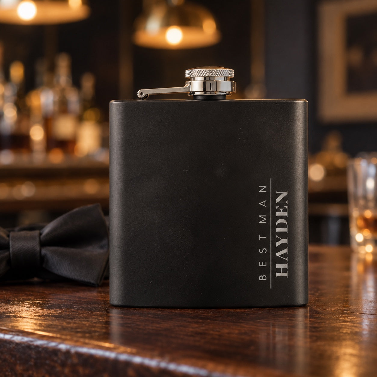 Engraved groomsmen flask in matte black on a walnut bar beside a black bow tie, the name HAYDEN running tall down the side with BEST MAN beside it