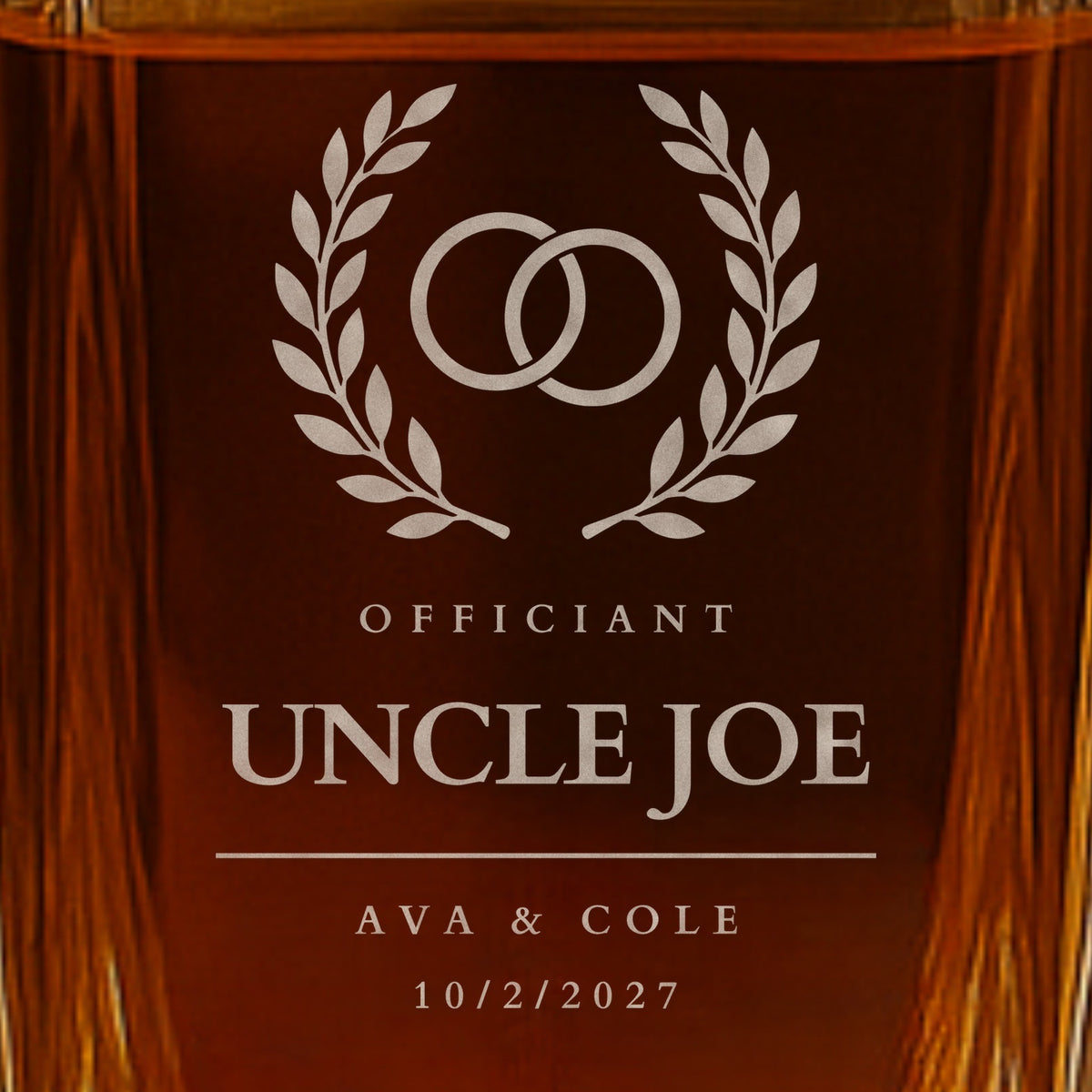 Close-up of frosted engraving on an officiant whiskey decanter: rings in a laurel wreath, engraved Uncle Joe with the couple Ava and Cole and 10/2/2027
