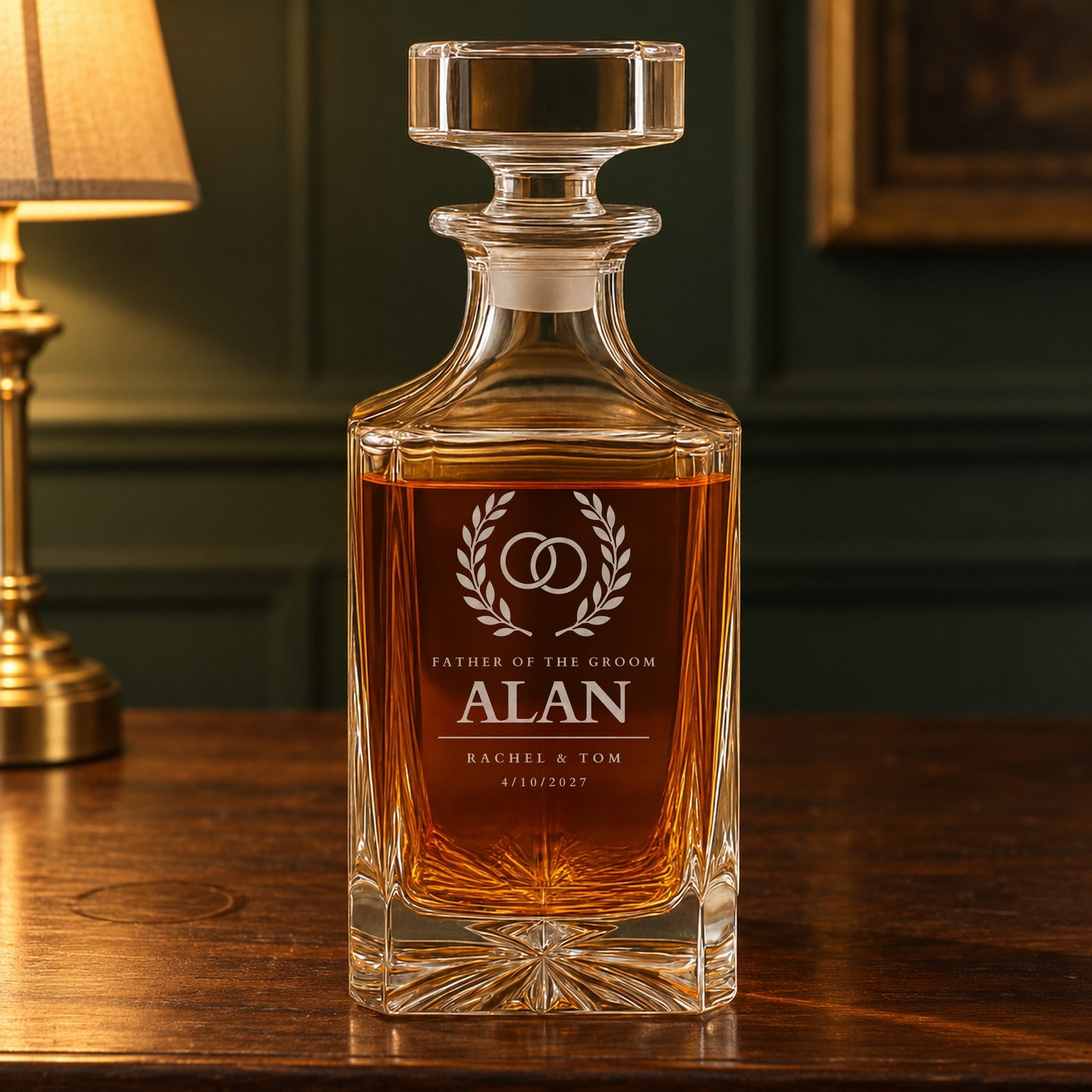 Front of a personalized father of the groom whiskey decanter by a brass lamp: rings in a laurel wreath, the name Alan, Rachel and Tom and the date 4/10/2027