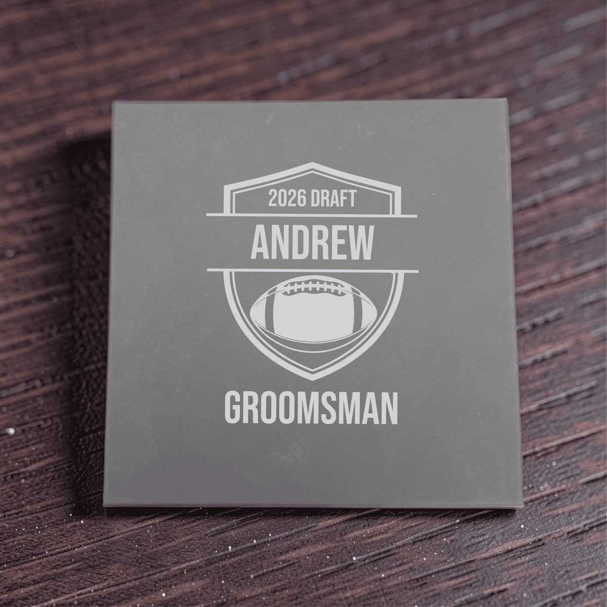 Gray gift box engraved with sample personalization reading "2026 DRAFT," "ANDREW," and "GROOMSMAN" with football shield design motif