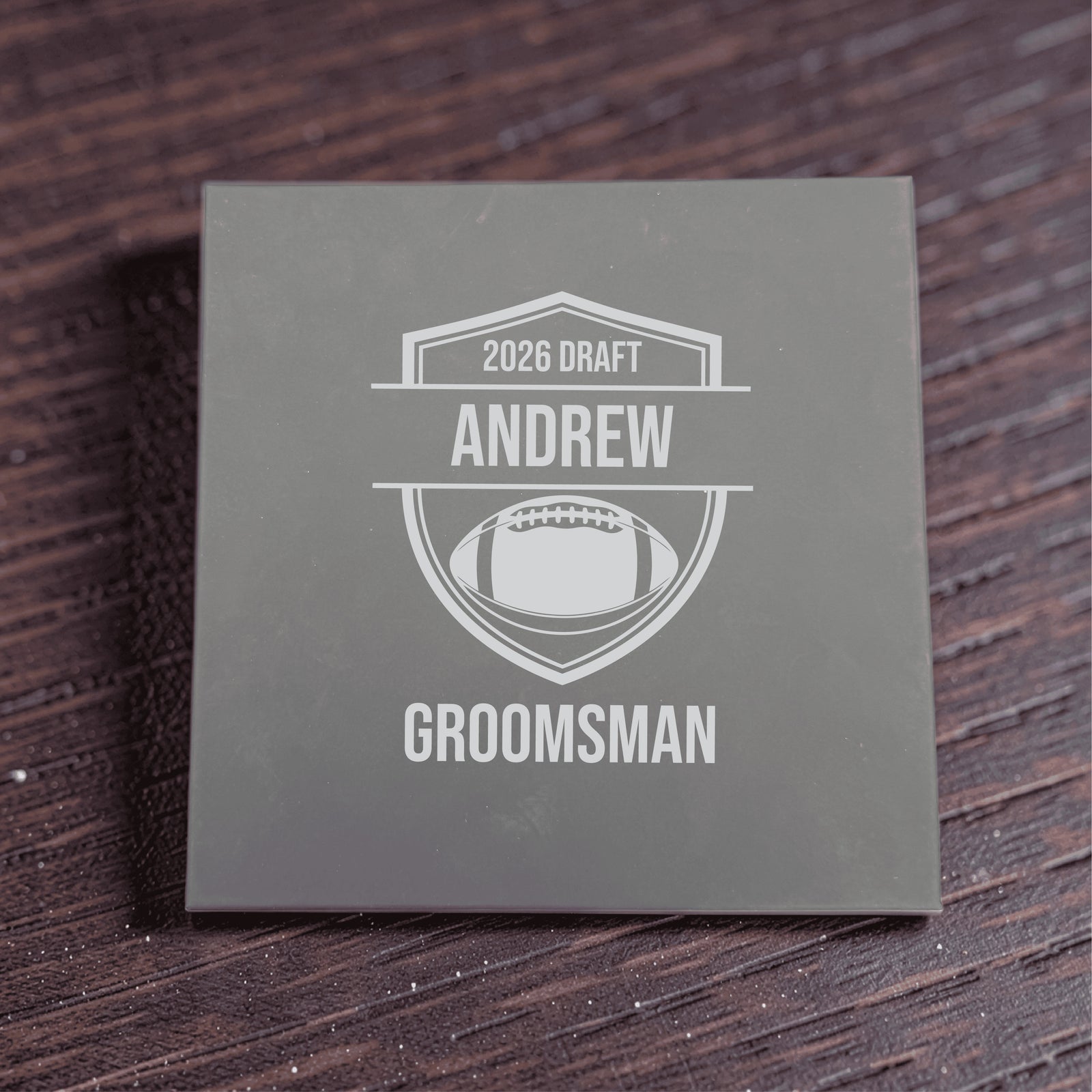 Gray gift box engraved with sample personalization reading "2026 DRAFT," "ANDREW," and "GROOMSMAN" with football shield design motif