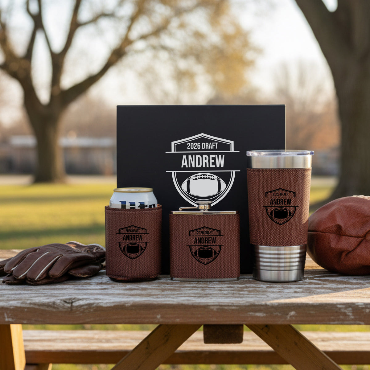 Brown leather koozies, can holder, and tumbler with brown gift box, all personalized with sample engraving "2026 DRAFT ANDREW" and football shield design, displayed outdoors on wooden table with football and gloves