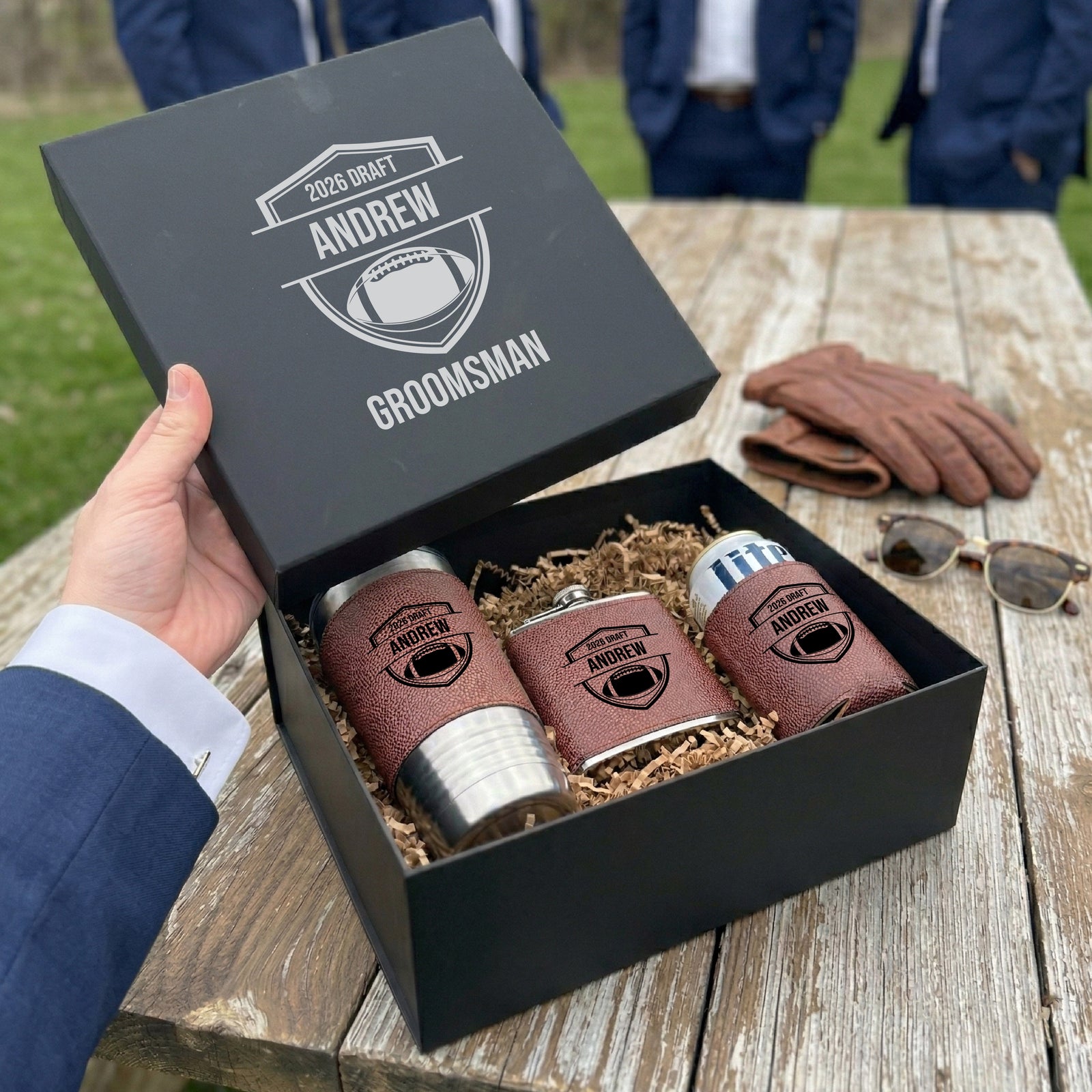 Charcoal gift box open to reveal brown leather flask, two matching koozies, and straw, all engraved with sample name "Andrew" and football motif design