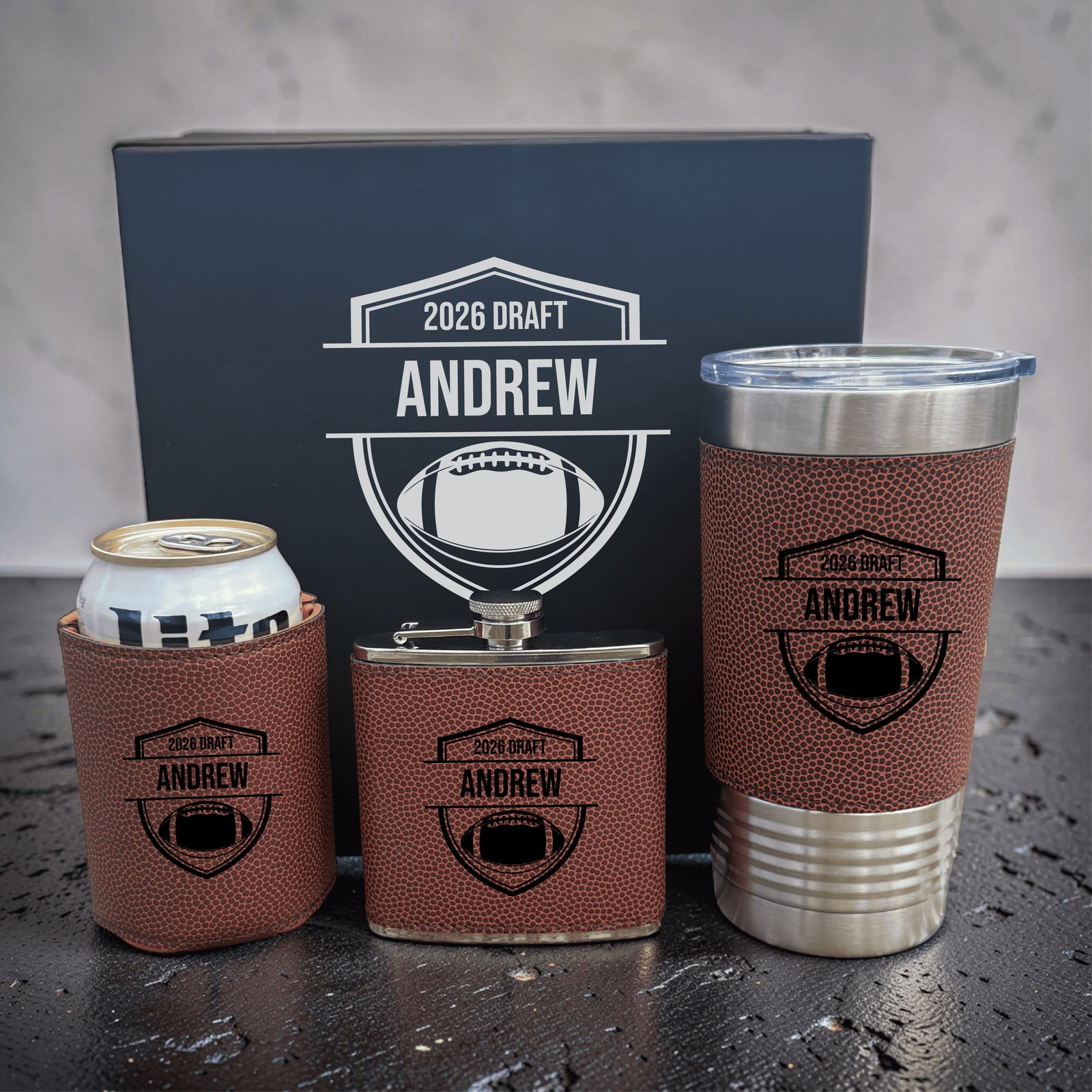 Three personalized koozies and tumbler with football-textured brown leather wraps engraved "2026 DRAFT ANDREW" with shield emblem, displayed with matching navy gift box in background.