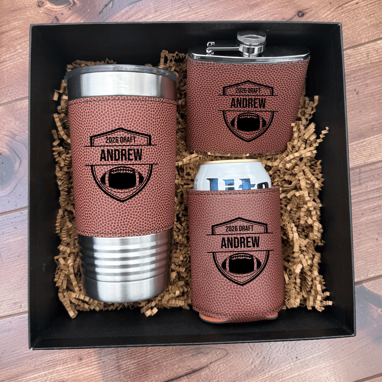 Three-piece gift set in black box with shredded kraft paper: insulated tumbler, stainless steel flask, and koozie, each with football texture and personalized sample engraving "2025 DRAFT ANDREW" with shield emblem.