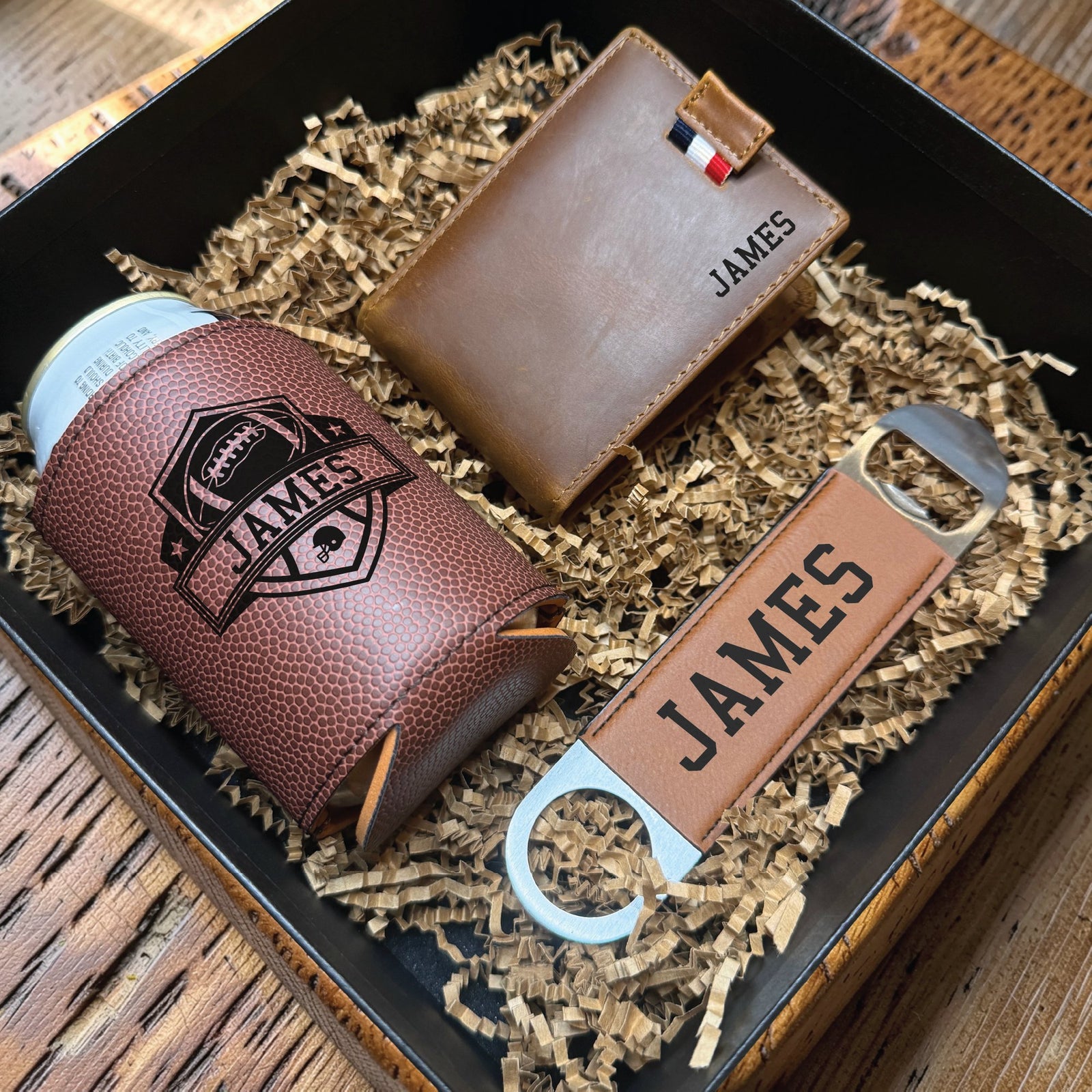 Football draft groomsmen proposal box open on a table, showing a football grain can cooler, leather bi-fold wallet and bar blade bottle opener each engraved with the name James.