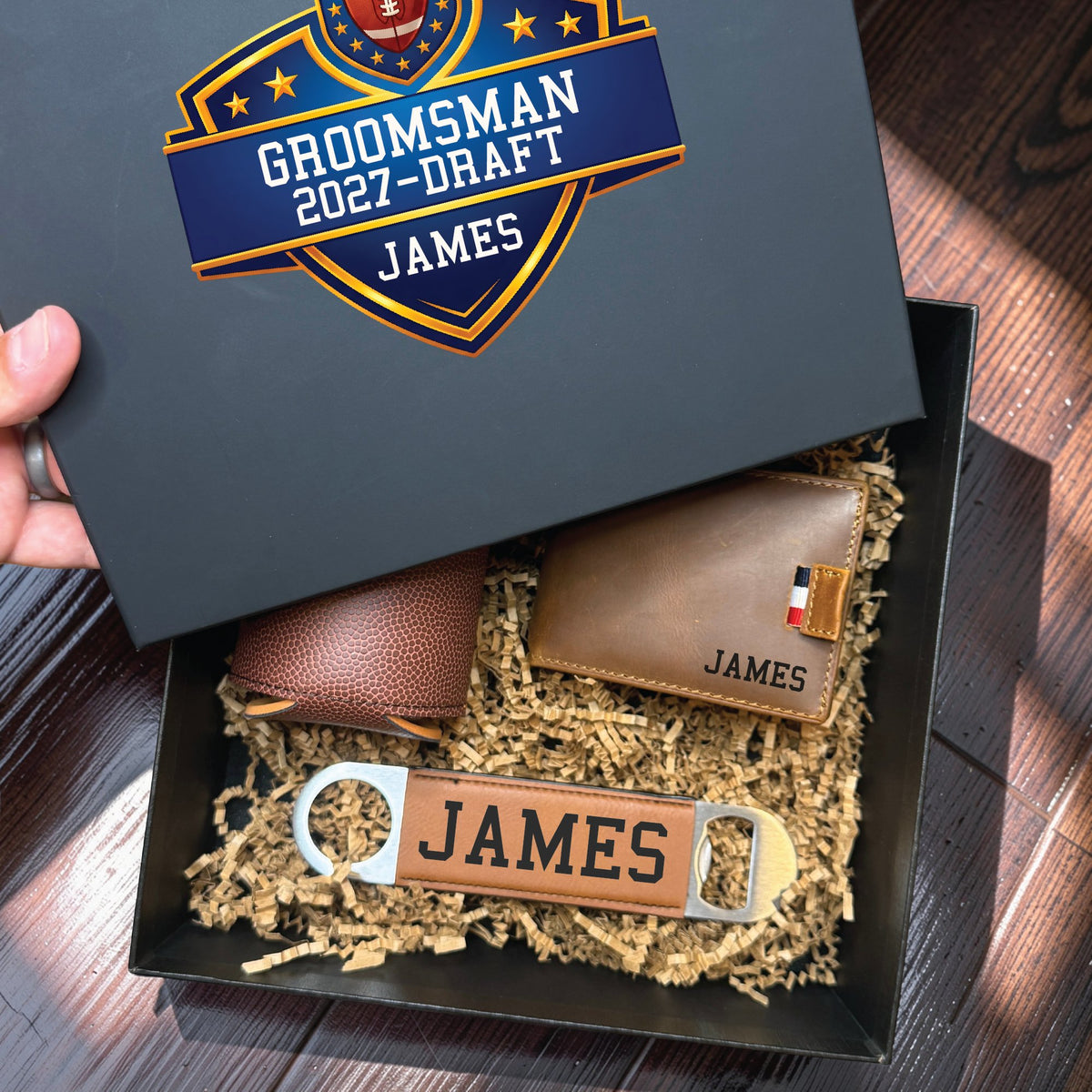 Hands lifting the printed lid off a groomsmen proposal box, revealing the football draft shield printed in colour with the role Groomsman and the name James.