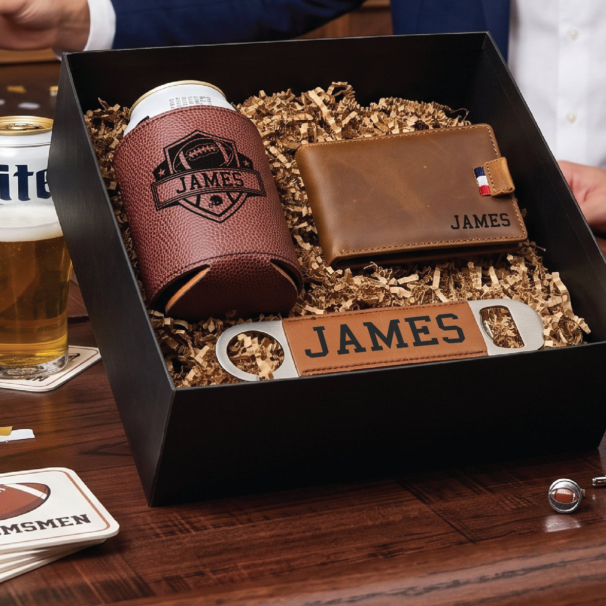 Open football draft groomsmen proposal box on a bar top beside a poured beer, with the engraved wallet, can cooler and leather wrapped bottle opener inside.