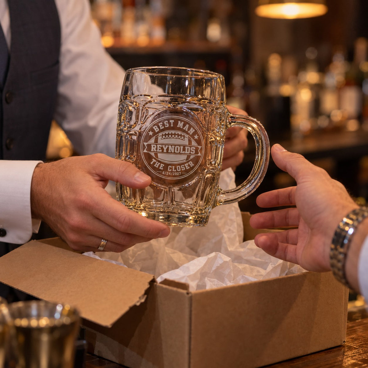 Groom handing an engraved football beer stein to his best man from a kraft gift box with tissue paper, personalized with Reynolds and The Closer