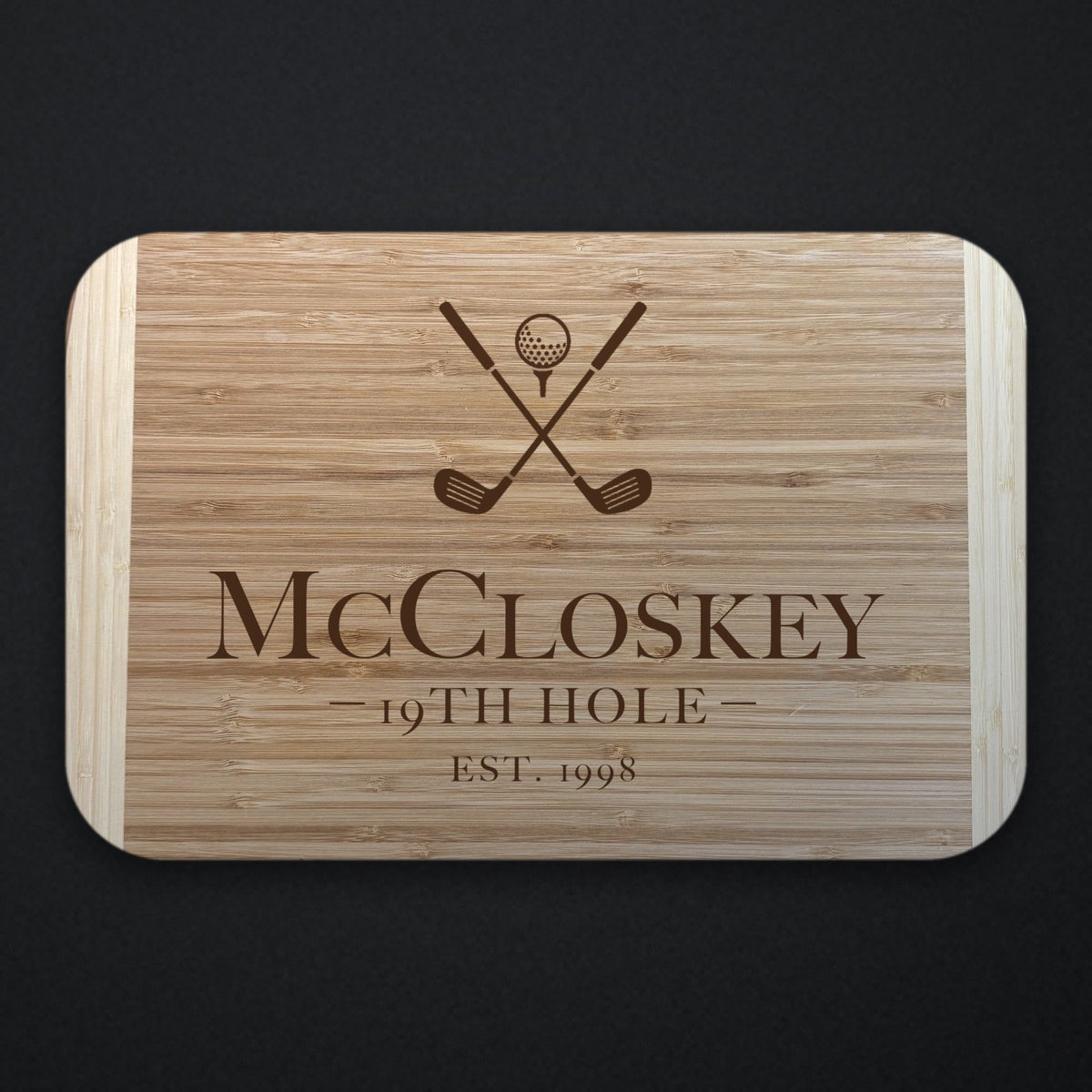 19th Hole golf groomsman cutting board in two tone bamboo engraved with crossed clubs and a ball above McCloskey, 19TH HOLE and EST. 1998, on a dark slate background.