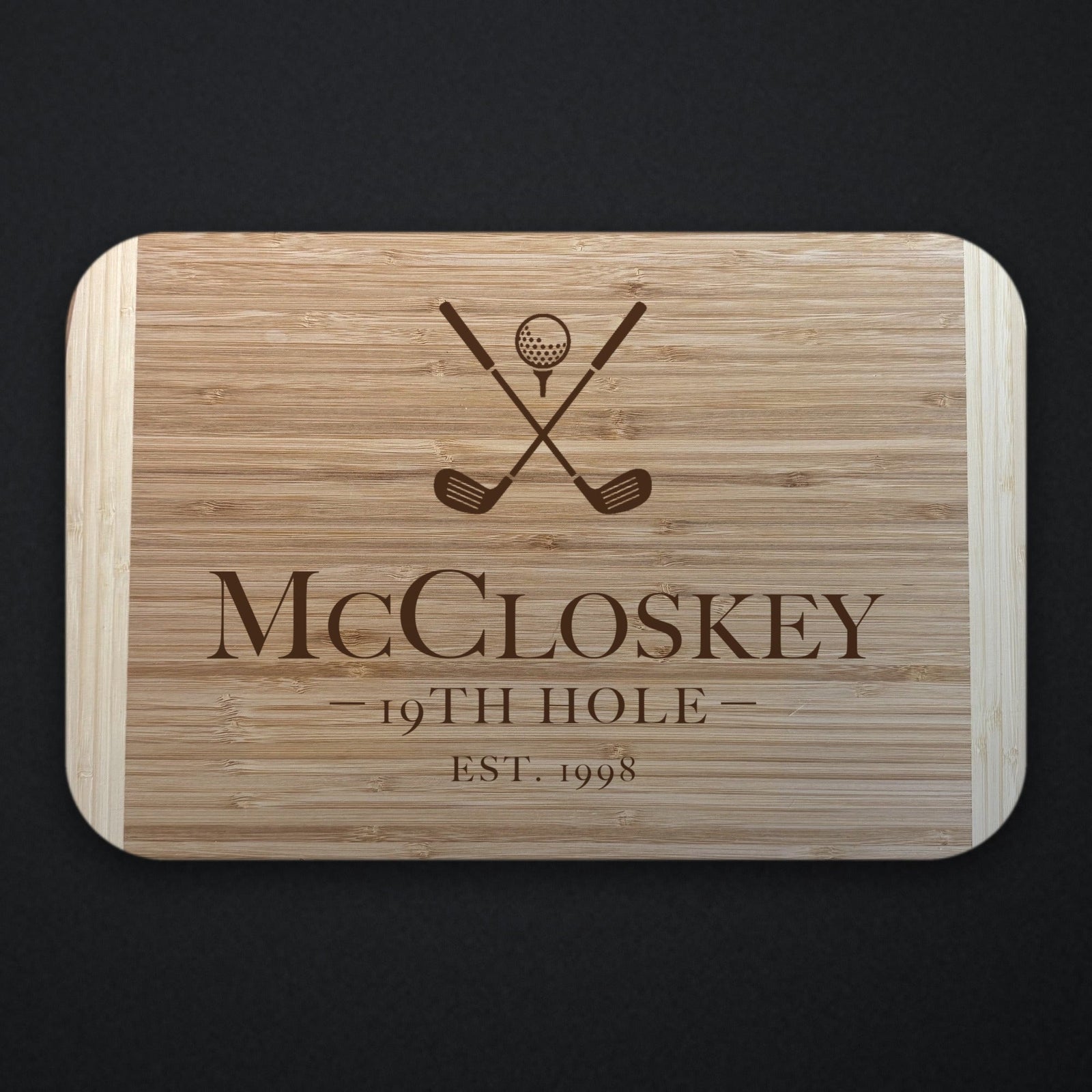 19th Hole golf groomsman cutting board in two tone bamboo engraved with crossed clubs and a ball above McCloskey, 19TH HOLE and EST. 1998, on a dark slate background.