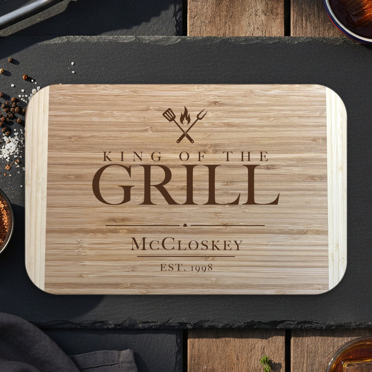 King of the Grill cutting board for dad on dark slate with grilling tongs, dry rub and bourbon, engraved McCloskey EST. 1998 under crossed grill tools on two tone bamboo.