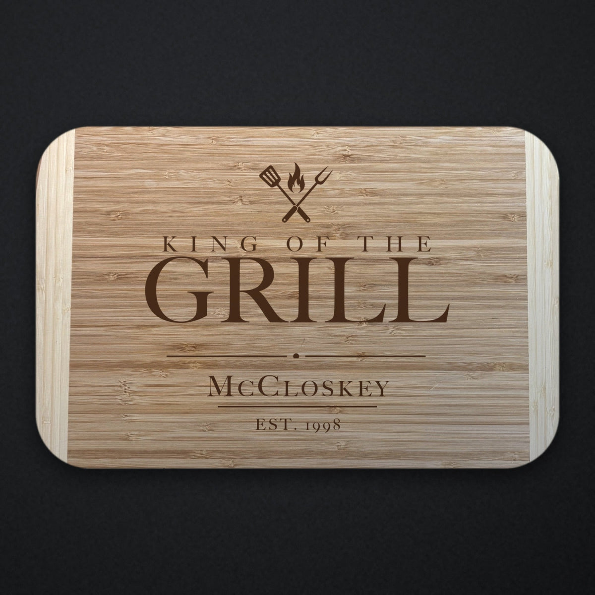 Father of the groom King of the Grill cutting board in two tone bamboo engraved under crossed grilling tools and a flame, with McCloskey and EST. 1998 below.