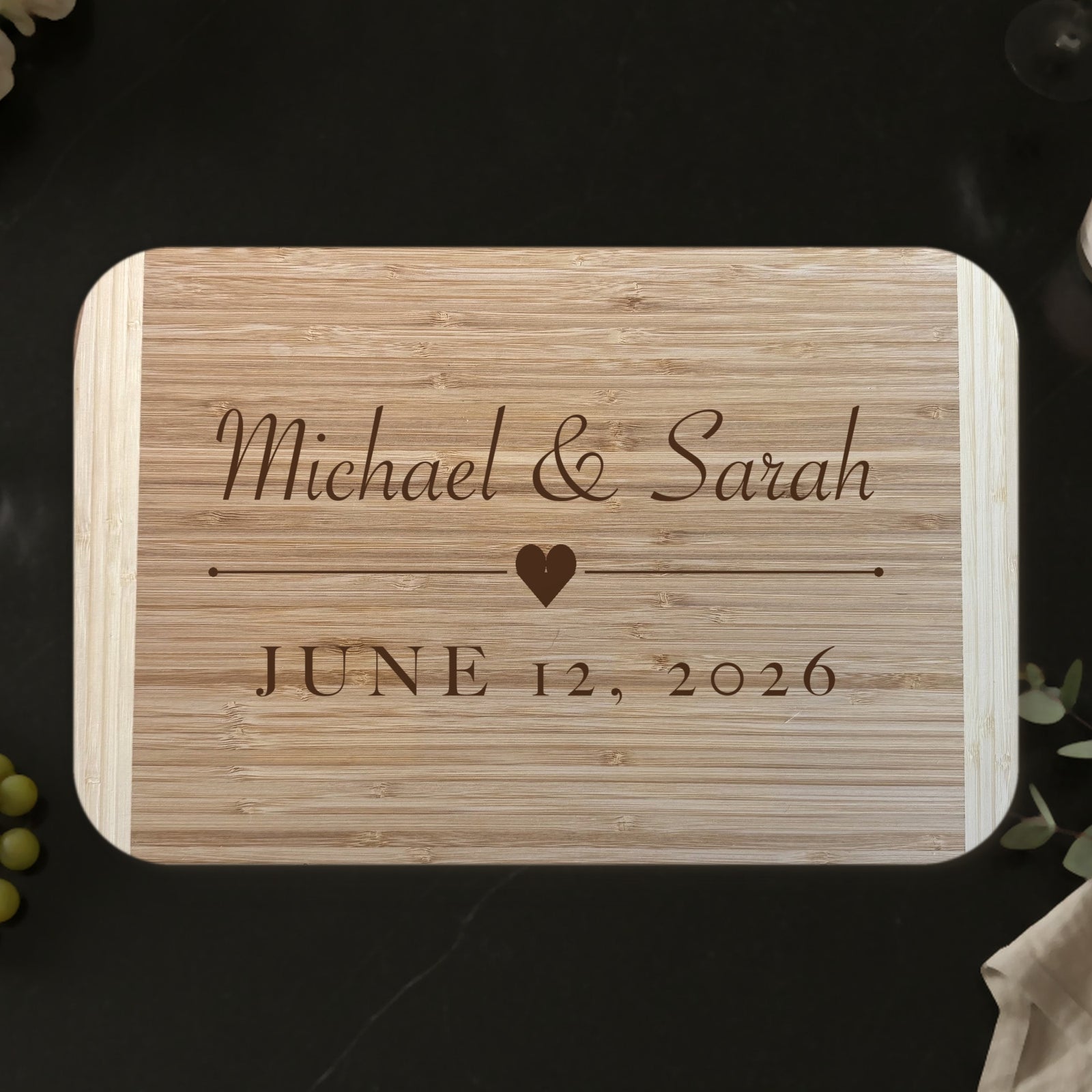 Wedding gift cutting board on a black marble countertop with roses and champagne, engraved Michael and Sarah above a heart with JUNE 12 2026 on two tone bamboo.