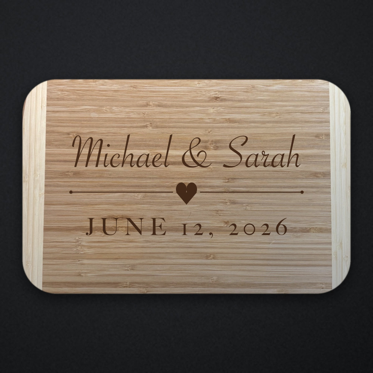 Two tone bamboo wedding cutting board engraved with Michael and Sarah in script above a heart and rule, with JUNE 12 2026 beneath, shown on a dark background.