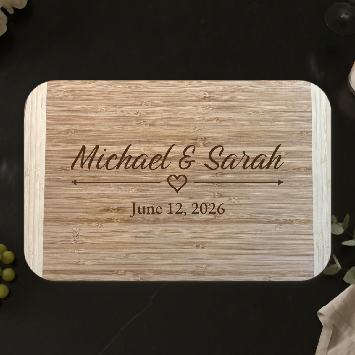 Wedding gift cutting board on a black marble countertop with roses and a glass of red wine, engraved Michael and Sarah in script above an outline heart and June 12 2026.