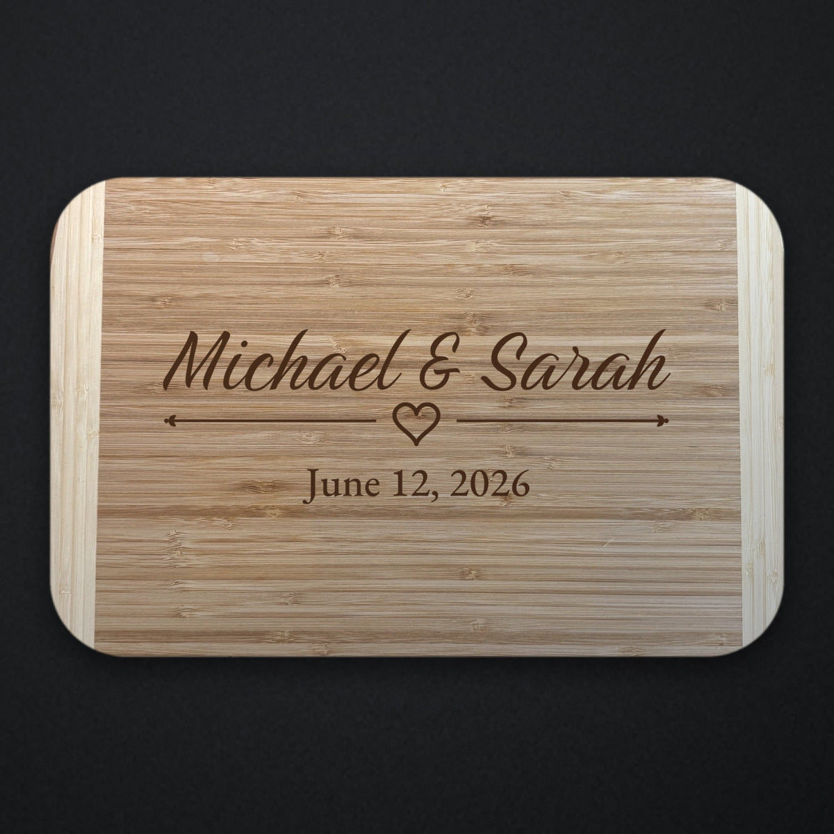 Two tone bamboo wedding cutting board engraved with Michael and Sarah in flowing script, an outline heart between two rules, and June 12 2026 underneath.