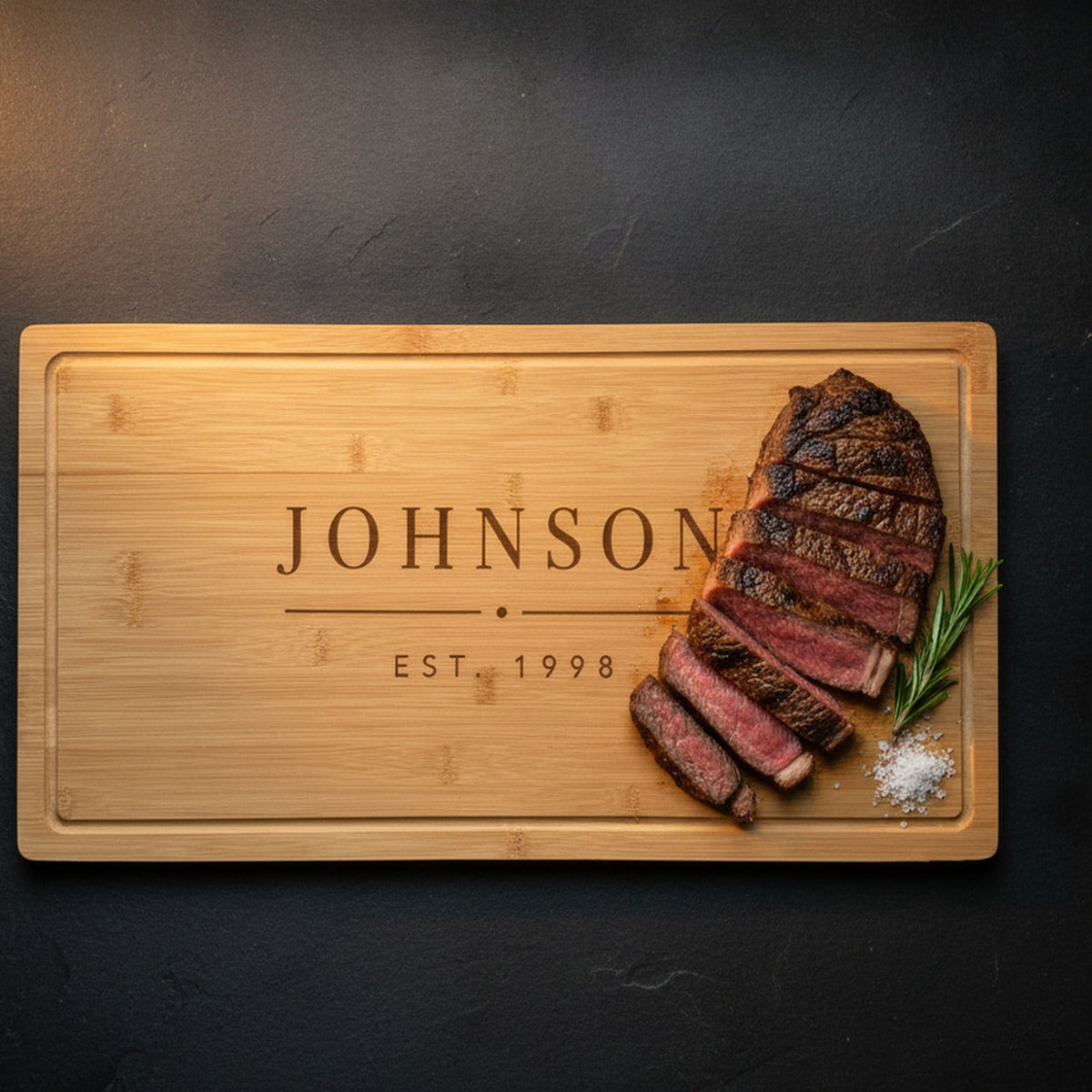 Engraved last name cutting board on dark slate with a sliced ribeye and rosemary resting on the bamboo, the family name JOHNSON and EST. 1998 beside it.