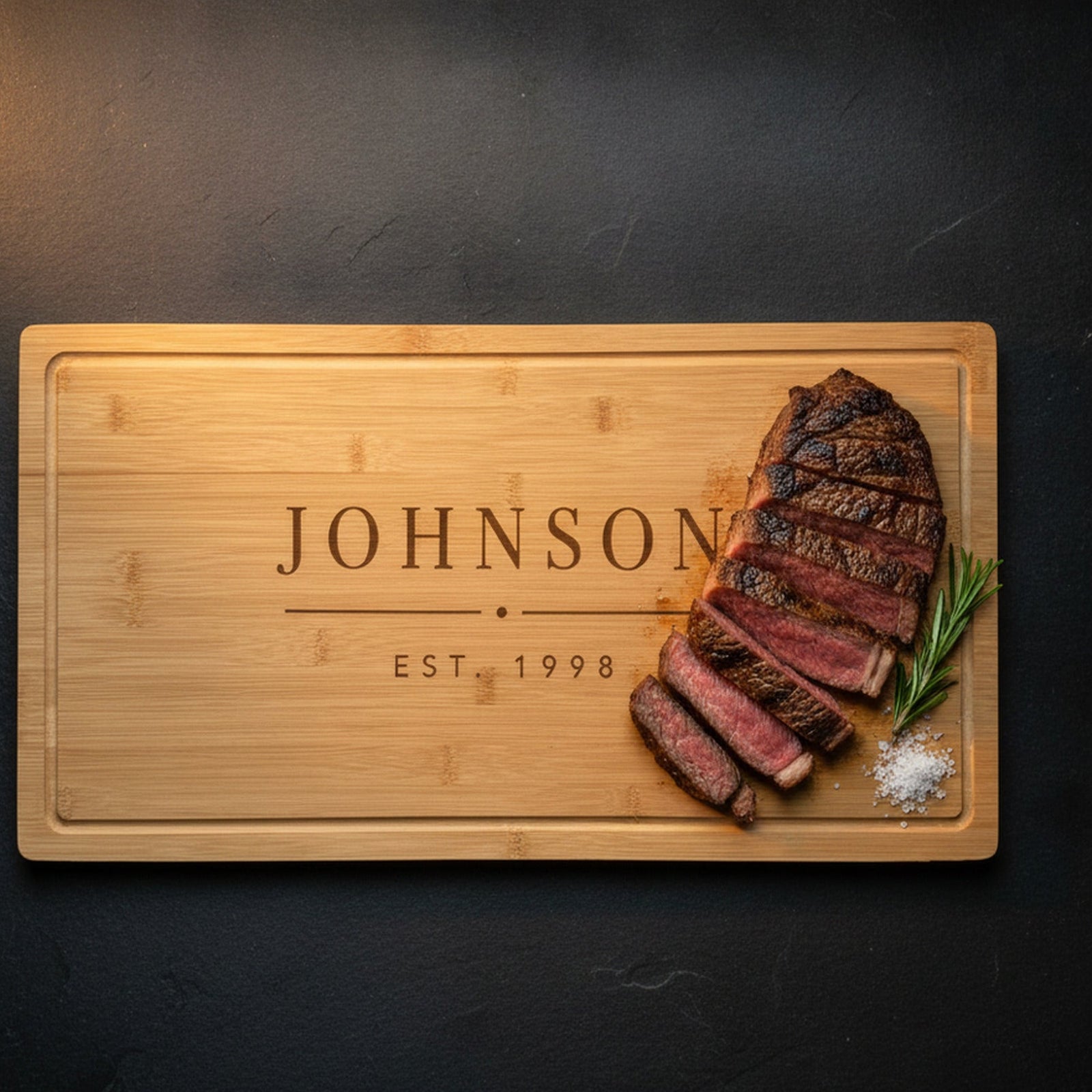 Engraved last name cutting board on dark slate with a sliced ribeye and rosemary resting on the bamboo, the family name JOHNSON and EST. 1998 beside it.