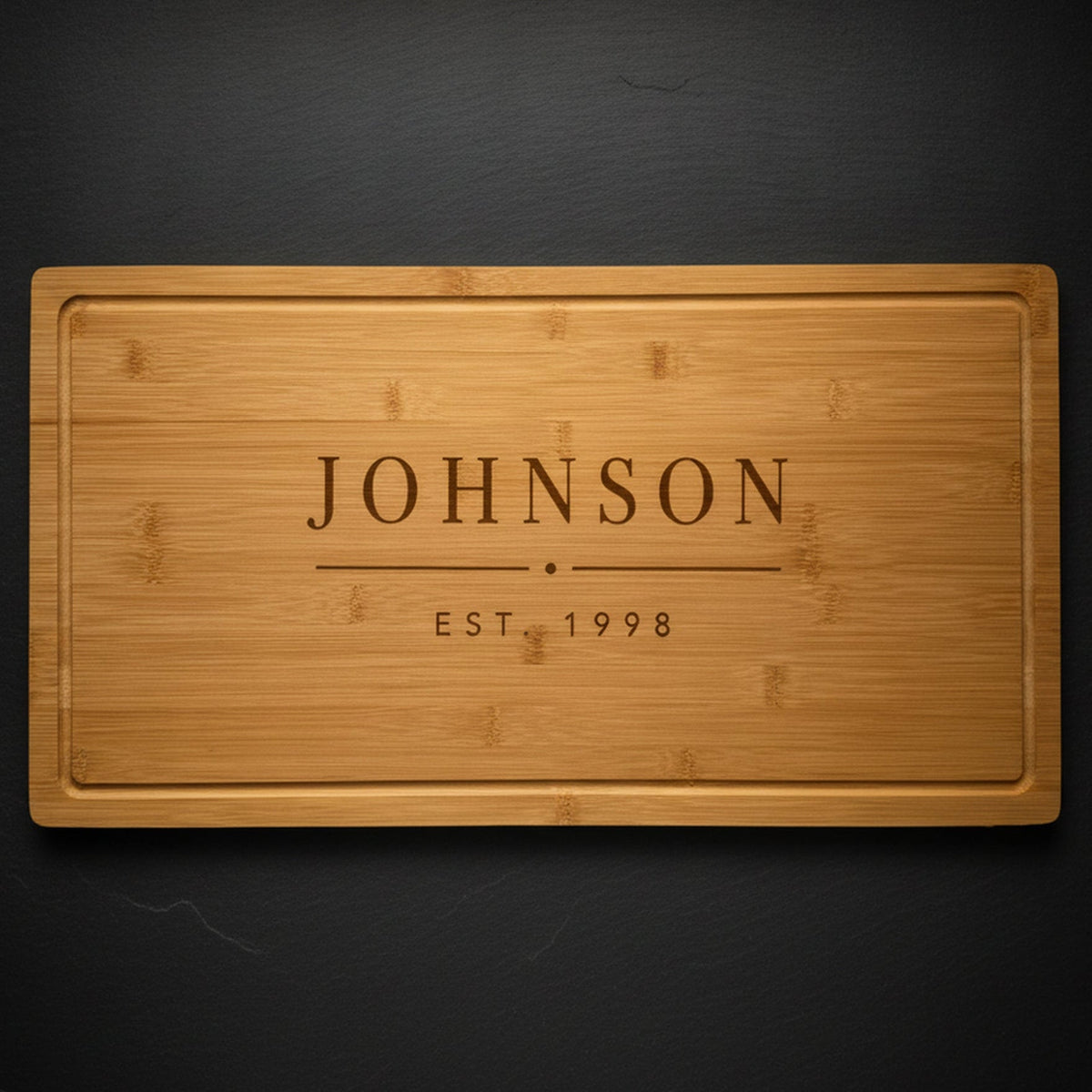 Personalized last name cutting board in solid bamboo on dark slate, engraved JOHNSON above EST. 1998 in clean block capitals, a wedding gift for the couple.