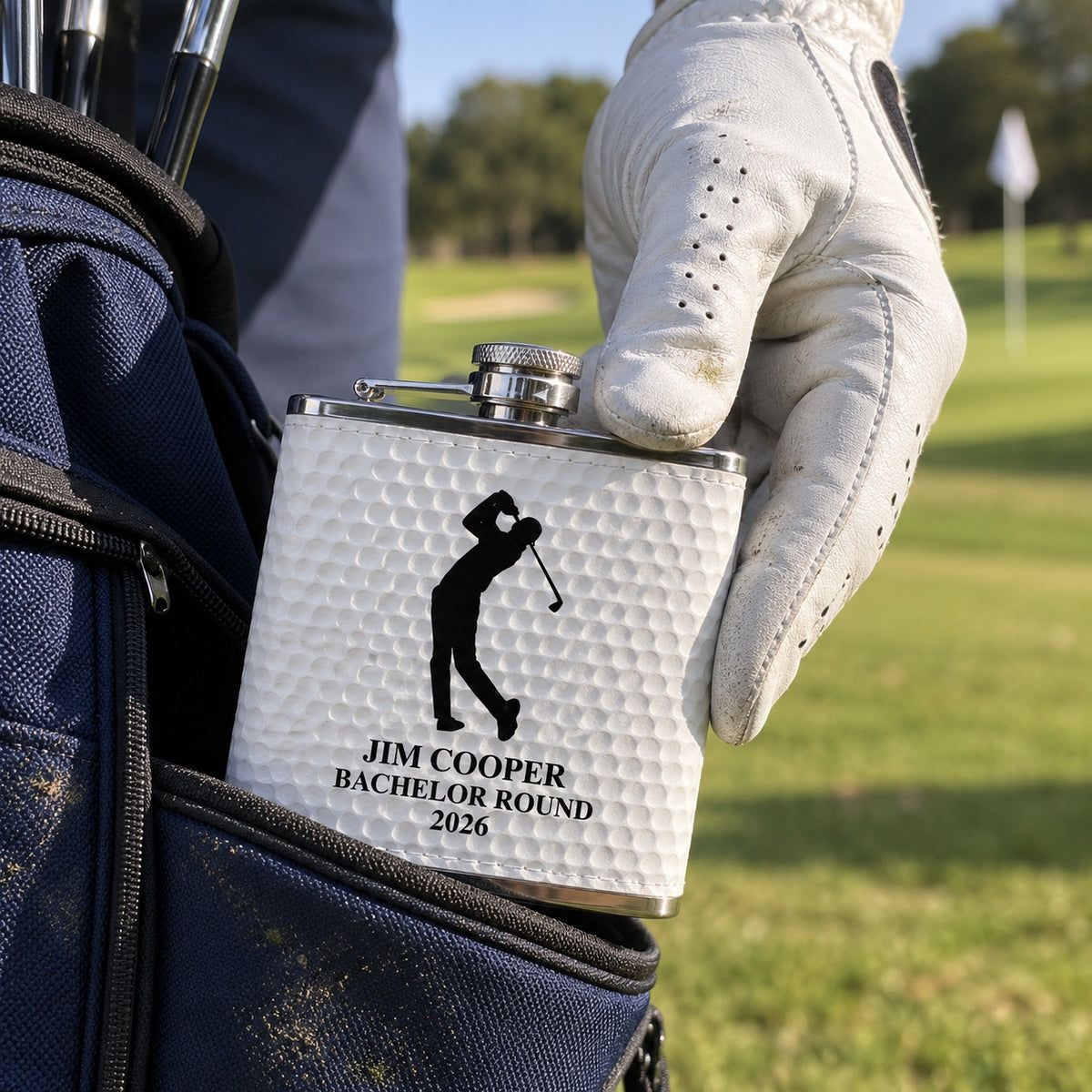 A golfer in a white glove pulls his golf ball flask engraved JIM COOPER BACHELOR ROUND 2026 out of his golf bag pocket on the course.