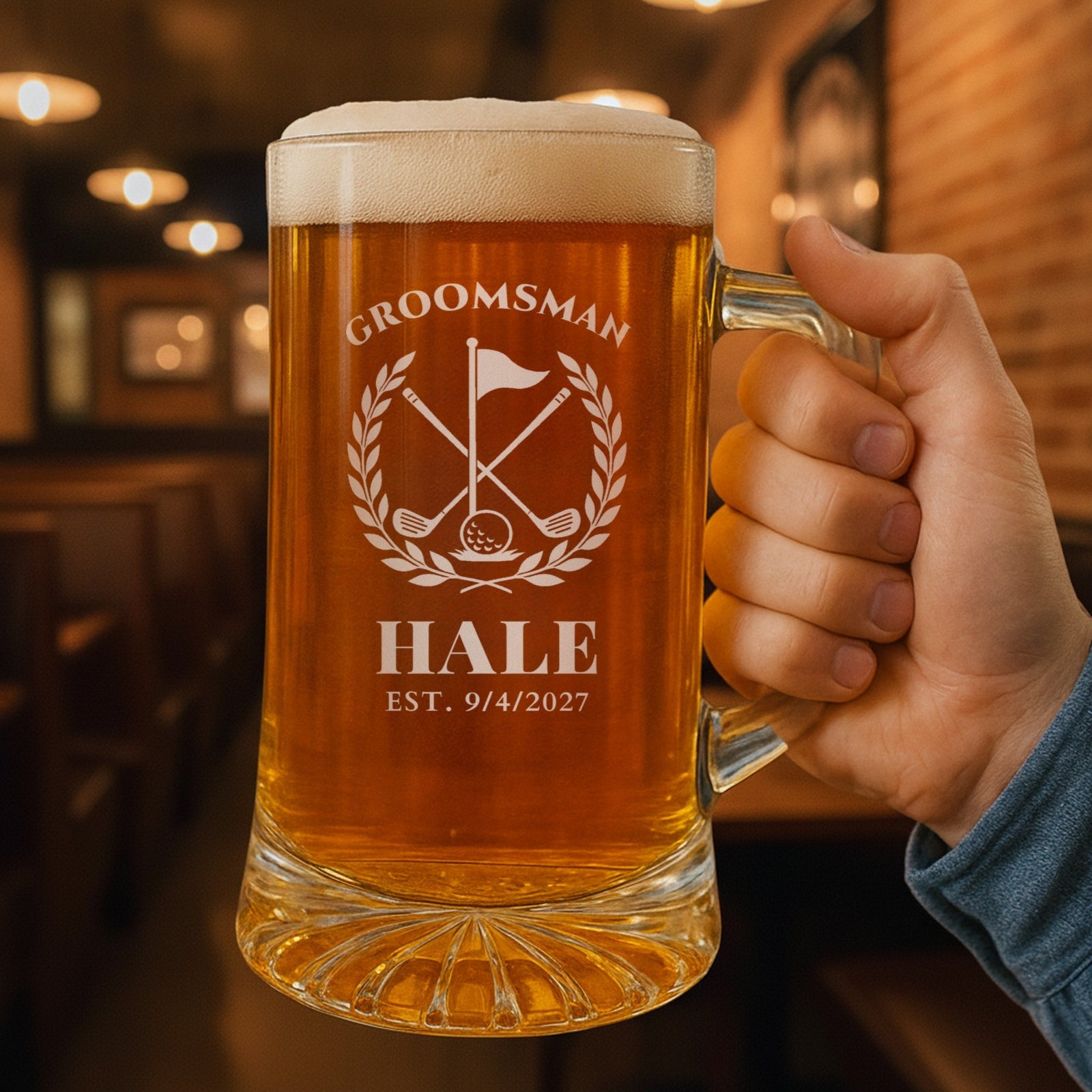 Hand holding the 25 oz personalized golf groomsmen beer stein in a pub, engraved with a golf flag, crossed clubs and laurel: Groomsman, Hale, Est. 9/4/2027
