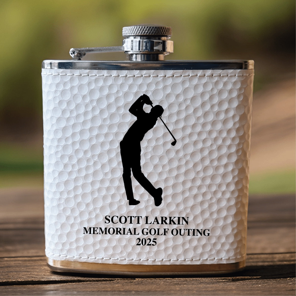 Real photo of the golf ball flask engraved with a golfer silhouette, SCOTT LARKIN MEMORIAL GOLF OUTING 2025, standing on a wooden table outdoors.
