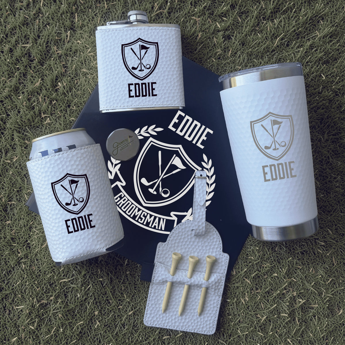 Groomsmen gift set with flask, tumbler, can cooler, and golf tees on grass.