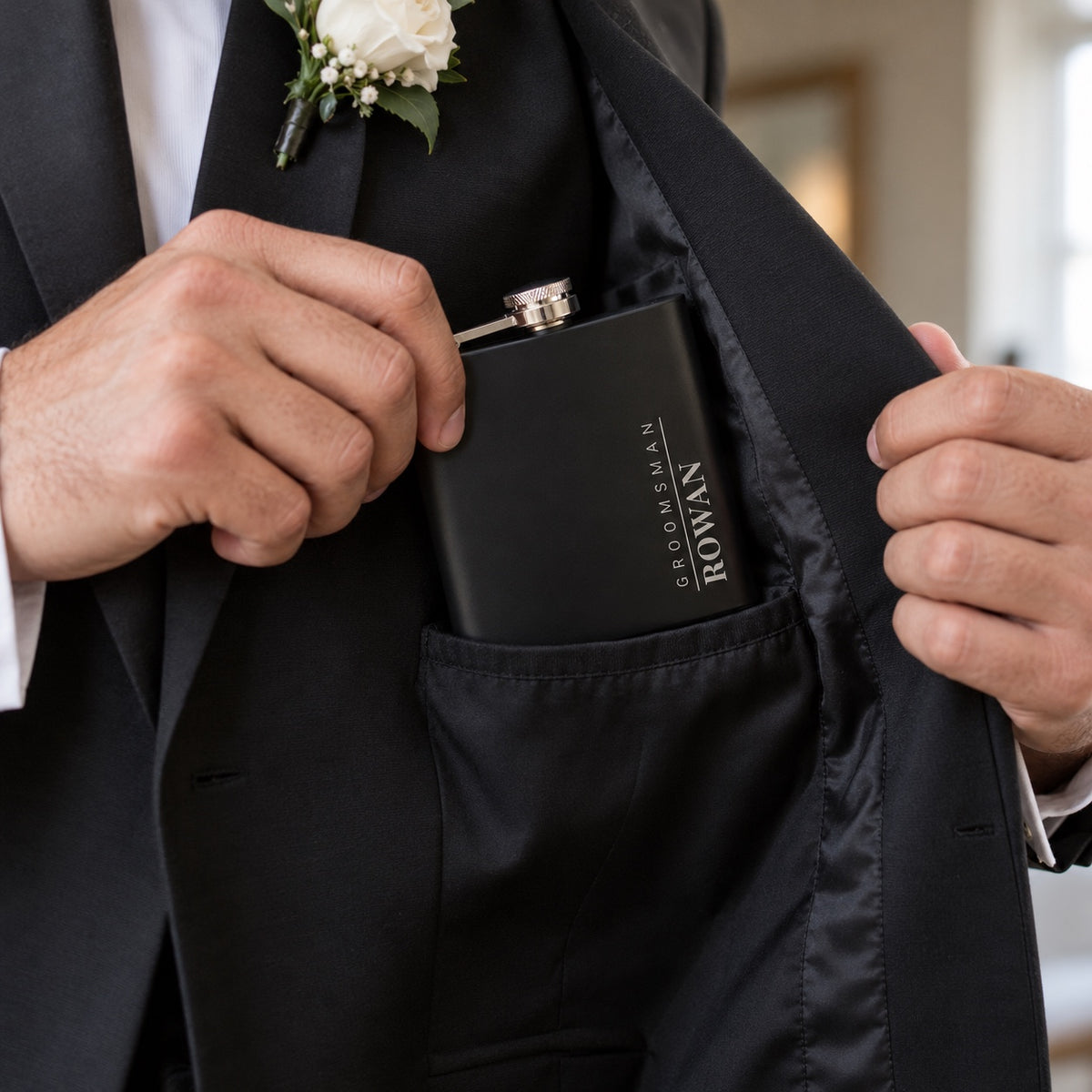 A groomsman slips his engraved flask, GROOMSMAN and ROWAN, into the inside pocket of his black suit jacket before the wedding, a white rose boutonniere on his lapel