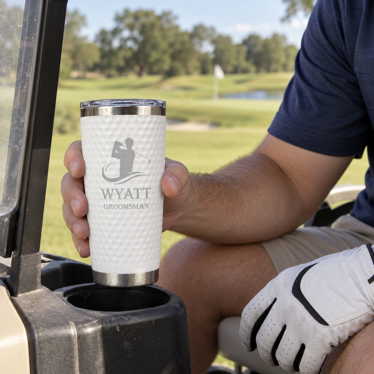 A groomsman lifts his golf ball tumbler engraved WYATT GROOMSMAN out of a golf cart cup holder during a bachelor party round on the course.