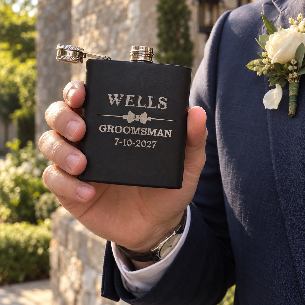 A groomsman in a navy suit and boutonniere holds his black flask engraved WELLS GROOMSMAN 7-10-2027 outside the venue before the wedding ceremony.