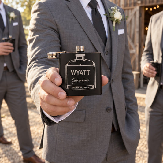 A groomsman in a grey suit holds out his black flask engraved WYATT, Groomsman and October 16, 2027 outside a barn venue before the wedding ceremony.