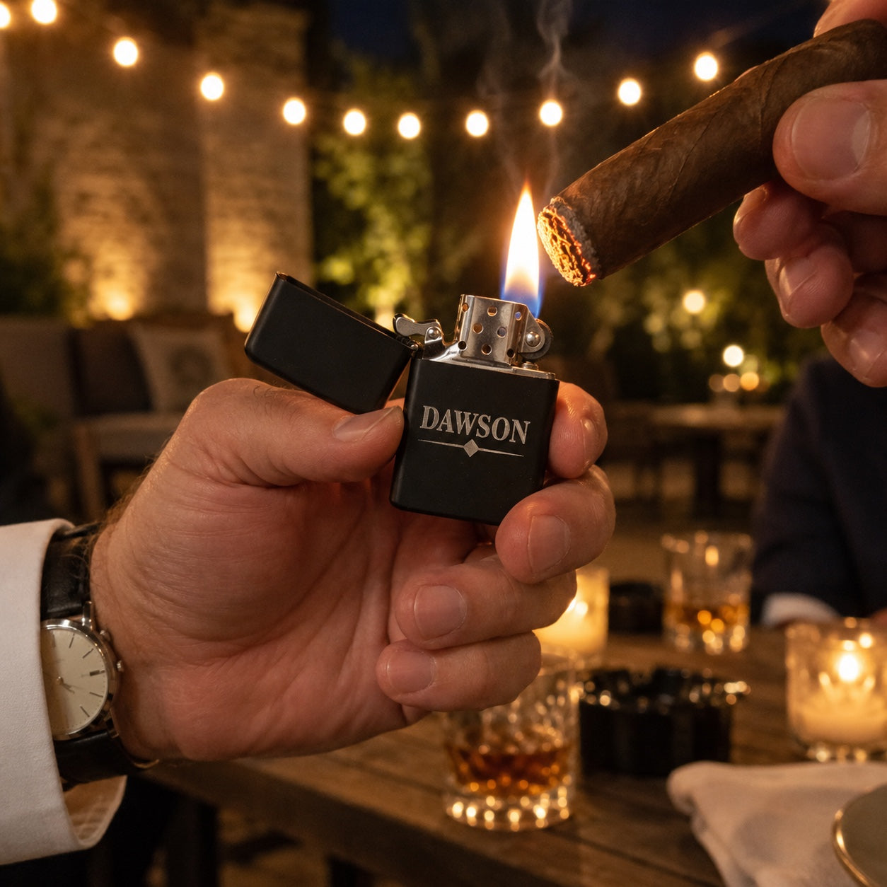 A groomsman lights a friend's cigar with his flip lighter engraved DAWSON on a string-lit patio after the rehearsal dinner, the flame steady.