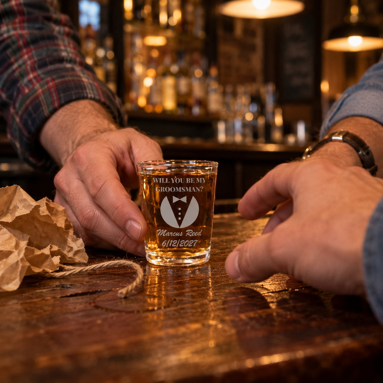 A man slides an engraved will you be my groomsman shot glass across a bar to his friend, Marcus Reed and a date engraved under the tuxedo, kraft paper beside it