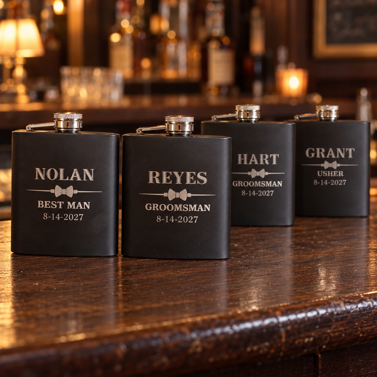Four black groomsmen flasks for the wedding party, engraved NOLAN Best Man, REYES and HART Groomsman and GRANT Usher, each with a bow tie and 8-14-2027.
