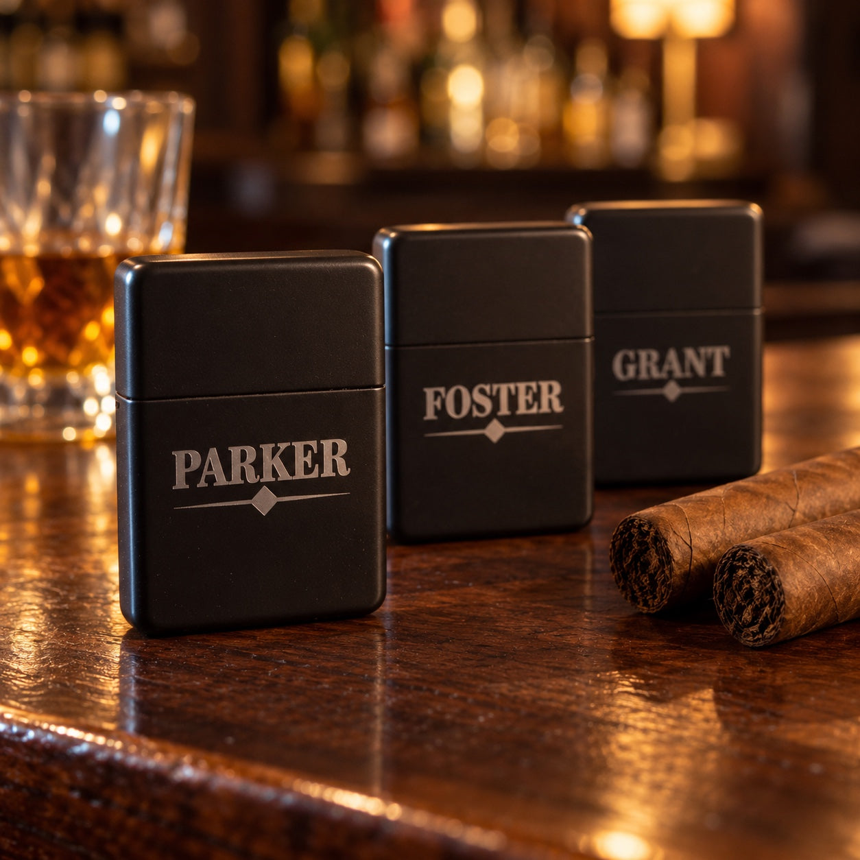 Three personalized cigar lighters for groomsmen in matte black, engraved PARKER, FOSTER and GRANT, standing beside two cigars on a dark wood bar.
