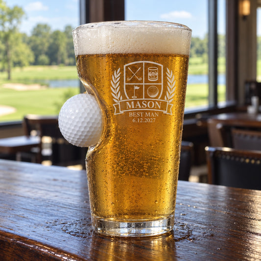 Groomsmen golf ball pint glass with a real golf ball set into its side, engraved with a golf crest, MASON, BEST MAN and 6.12.2027, on a clubhouse bar.