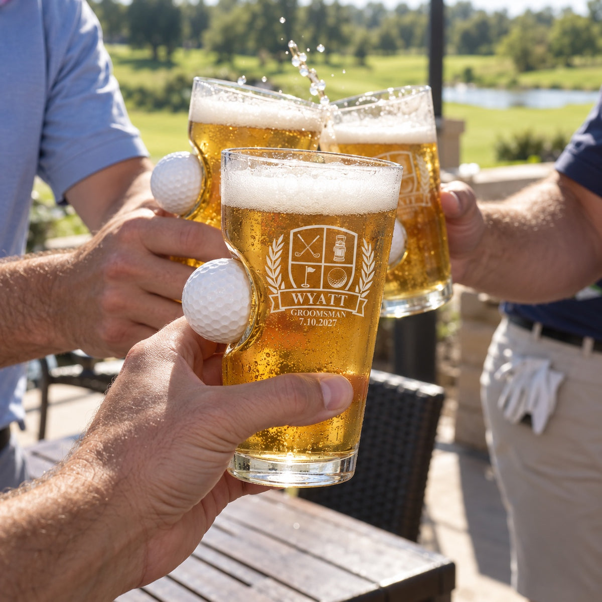 Groomsmen toast with golf ball pint glasses on a clubhouse patio after a bachelor party round, the front glass engraved WYATT GROOMSMAN 7.10.2027.