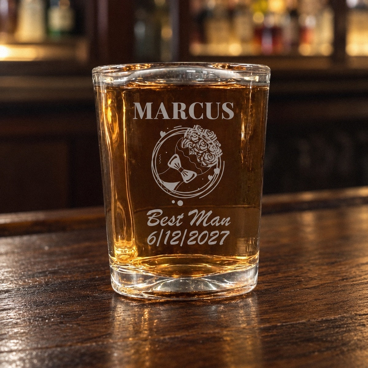 Groomsmen shot glass with whiskey on a walnut bar, engraved MARCUS over a floral crest and bow tie design with Best Man and 6/12/2027