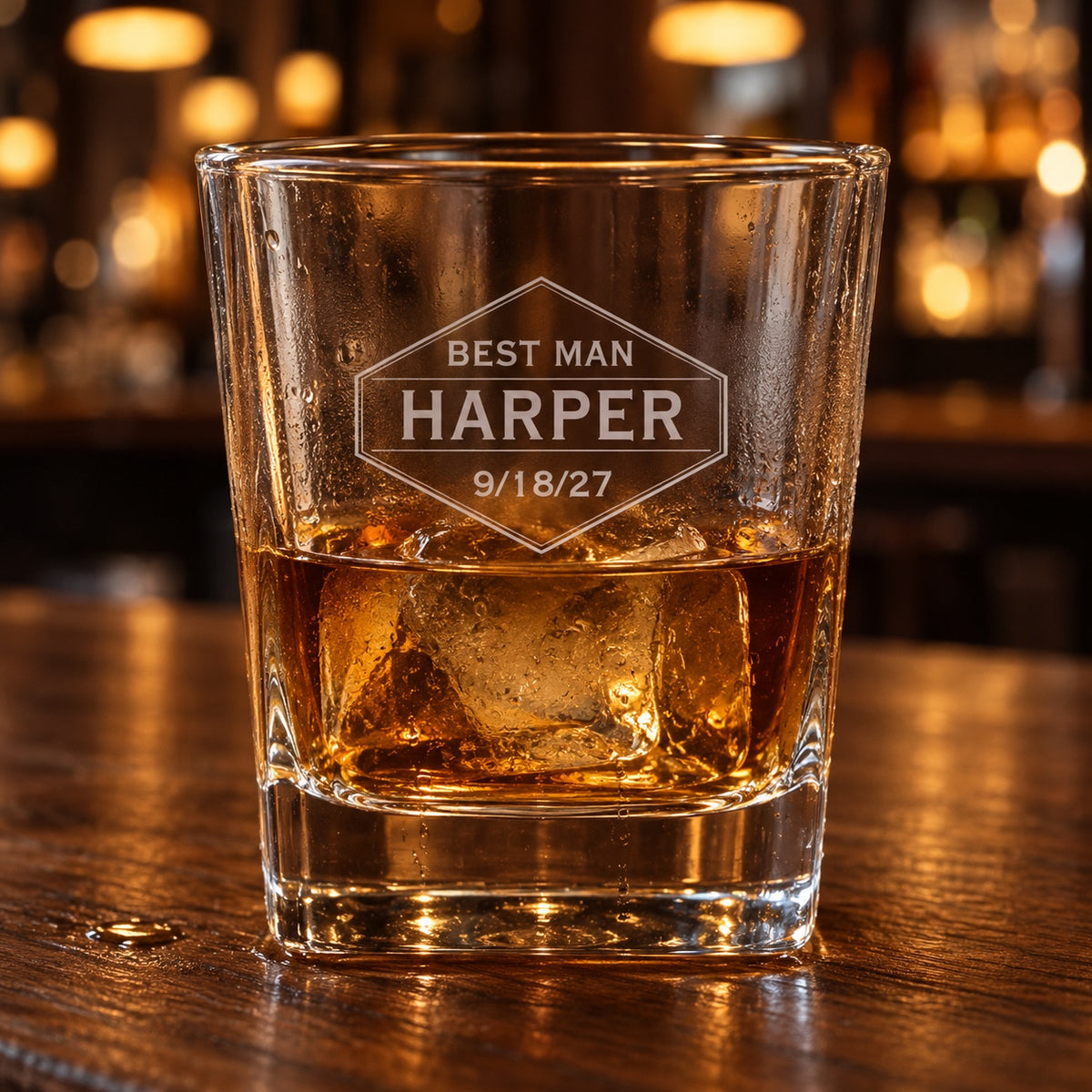 Personalized groomsmen whiskey glass engraved in a hexagon badge with BEST MAN, HARPER and 9/18/27, holding whiskey over a large ice cube on a walnut bar.